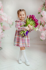 Cute adorable little girl in a dress holding a bouquet of tulip flowers, in a bright photo studio.