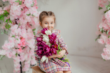 Fototapeta premium Cute adorable little girl in a dress holding a bouquet of tulip flowers, in a bright photo studio.