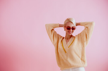 Happy smiling woman wearing trendy pink sunglasses, yellow v-neck sweater, posing on pink...