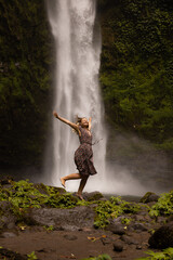 Fototapeta na wymiar Happy Caucasian woman jumping near the waterfall. Legs in motion movement. Nature and environment concept. Travel lifestyle. Woman wearing dress. Copy space. Nung Nung waterfall in Bali