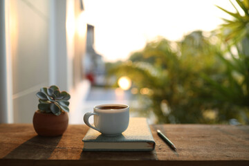 Obraz premium Coffee cup with small plant pot and notebook on wooden table under sunlight