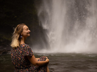 Young Caucasian woman sitting on the rock and enjoying waterfall landscape. Woman portrait. Energy of water. Travel lifestyle. Nung Nung waterfall, Bali