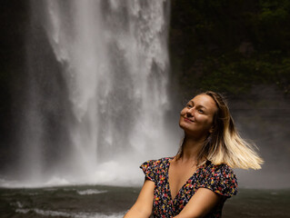 Young Caucasian woman sitting on the rock and enjoying waterfall landscape. Woman portrait. Energy of water. Travel lifestyle. Nung Nung waterfall, Bali