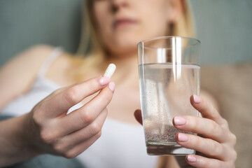 Stressed woman drinking white round pill while sitting in bed with glass of water in hand.