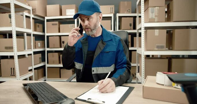 Senior warehouse worker in blue uniform sits at desk talking on mobile phone and looking at computer solves work problems.