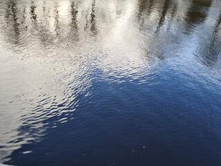 blurred reflections of trees, blue sky and white clouds on river surface with ripples because of wind
