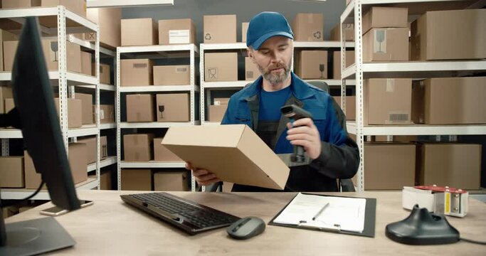 Senior warehouse worker in blue uniform sits at desk against background of cardboard boxes, scans parcel and looks for information about it on computer.