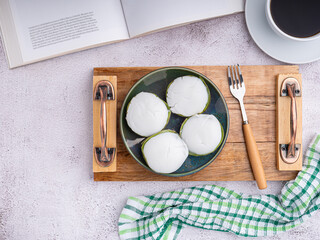 Top view of Thai pudding with coconut topping on a plate on a wooden tray with a book, a white coffee cup, and a cloth placed on a gray stone background. Space for text. Concept of relaxation