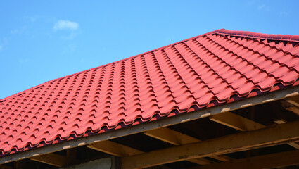 A close-up of an unfinished metal roof with red metal roofing tiles, wood ceiling joists, eaves, and roof beams.