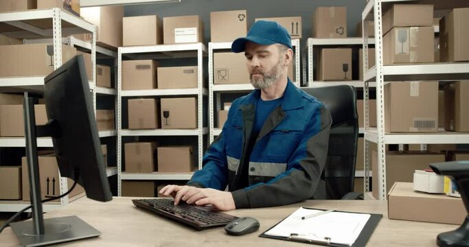 Senior warehouse worker in uniform sits in office at desk and works intently at computer typing on keyboard and searching for information.