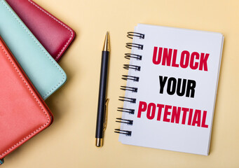 Multi-colored diaries lie on a beige background next to a pen and a notebook with the words UNLOCK YOUR POTENTIAL. Flat lay.