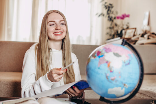 Caucasian girl student smiles, holds a notebook in his hands and studies geography on a globe while sitting at home in quarantine. Education abroad. Distance learning