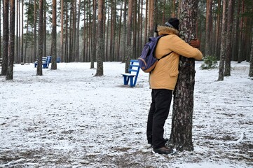 A man hugs a tree in a pine park forest in winter. Benches and snow. love nature