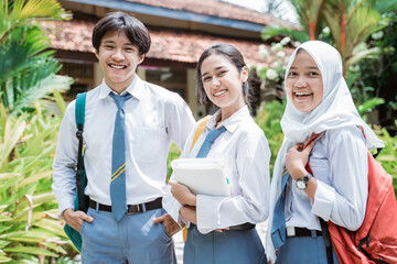 close up of a male Indonesian high school student and two female high school students wearing a school bag and smiling with the camera with ornamental plants and the school building as the background.