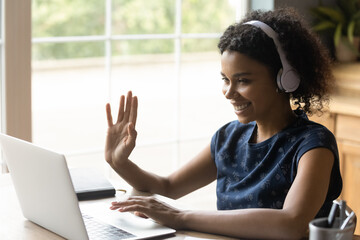 Ready to start. Positive biracial teen female in headset sit by laptop waving hand to camera greet teacher tutor at distant video lesson. Young black woman student learning online via web conference - obrazy, fototapety, plakaty