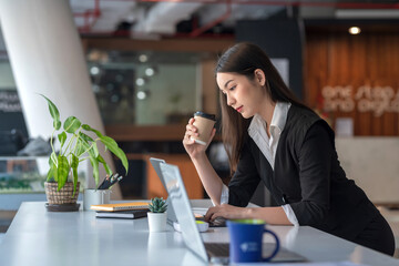 Side view beautiful Asian businesswoman holding a coffee mug working on a tablet at a modern office.