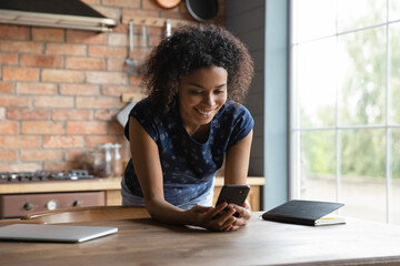 In a good mood. Happy young biracial woman stand by table near large kitchen window texting messaging online using cell device. Smiling black teen female spend time surfing web on modern smartphone