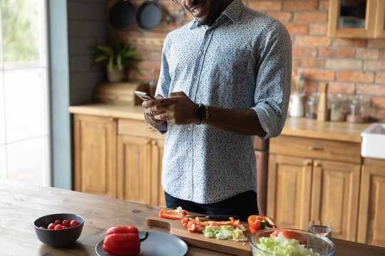 Check Web For Recipe. Cropped Shot Of Young African Male Stand By Kitchen Table Use Cell Search Details Of Preparing Dish Online. Black Man Husband Cooking Food Consult With Wife Using Wifi Internet