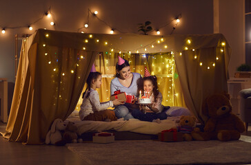 Obraz na płótnie Canvas Family time. Two twin sisters with their mother are sitting in a cozy tent bed and celebrating their birthday. Family in holiday hats exchanges gifts and gathers to blow out the candles on the cake.