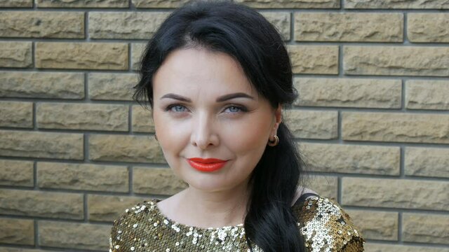 Beautiful spectacular young woman with stunning evening make-up smoky eyes with red lipstick in a sparkling gold dress with sequins on the street against the background of a brick brown wall