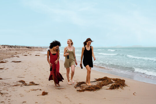 Delighted multiethnic women with beer walking on beach
