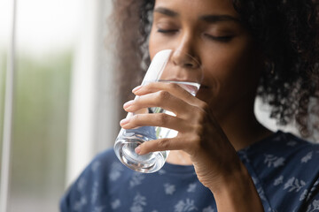 Closeup of young african woman drinking cool sparkling mineral water from glass hydrate having...