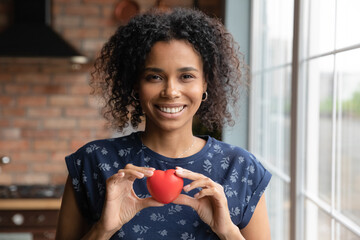 Headshot portrait of happy millennial black woman looking at camera holding small red toy heart in hands at breast height. Smiling young mixed race female express gratitude appreciation on photo video