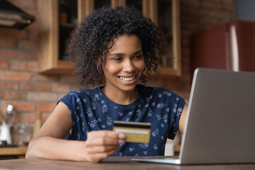 Happy biracial female vip bank customer pay online buy goods at internet shop using gold card for web payment. Smiling successful young black woman look at laptop screen check balance in e-banking app