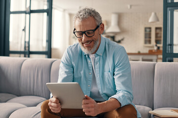 Cheerful senior man in casual clothing using digital tablet while sitting on the sofa at home - obrazy, fototapety, plakaty