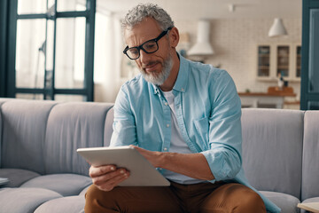 Concentrated senior man in casual clothing using digital tablet while sitting on the sofa at home