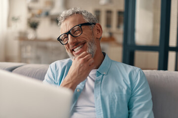 Cheerful senior man in casual clothing and eyeglasses using laptop and smiling while sitting on the sofa at home - obrazy, fototapety, plakaty