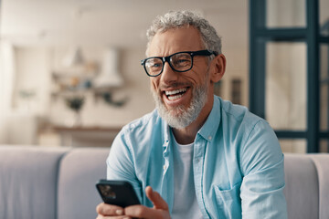 Cheerful senior man in casual clothing and eyeglasses using smart phone while sitting on the sofa...