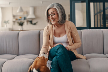 Obraz premium Happy senior woman in casual clothing spending time with her dog while sitting on the sofa at home