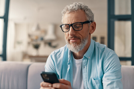 Senior man in casual clothing and eyeglasses texting someone using smart phone while sitting on the sofa at home