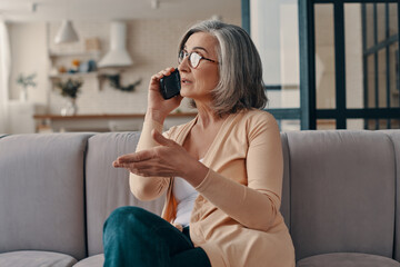 Beautiful senior woman in casual clothing talking on the smart phone while sitting on the sofa at home
