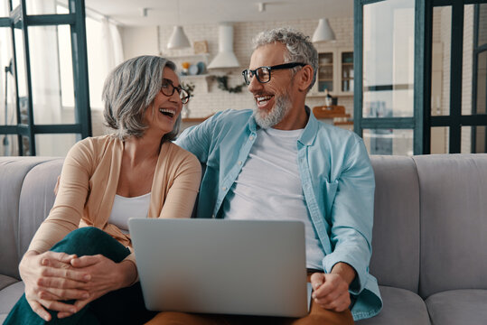 Happy senior couple in casual clothing smiling and using laptop while bonding together at home
