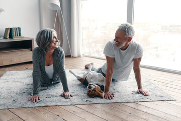 Loving senior couple in sports clothing doing yoga and smiling while spending time at home with...