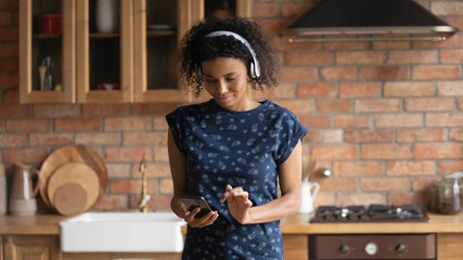 Young mixed race woman housewife wearing earphones use mobile phone app at home kitchen to find recipe online. Focused black female listen to music when cooking order fresh food products in internet
