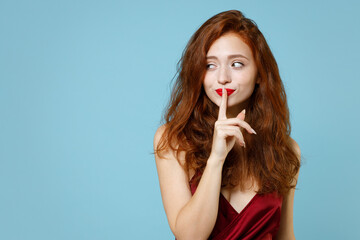 Young readhead ginger curly secret caucasian woman 20s in red party evening dress gown say hush be quiet with finger on lips shhh gesture look aside isolated on pastel blue background studio portrait.