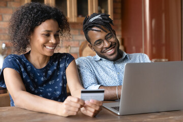 Paying online. Smiling young married couple of afro american ethnicity provide payment via electronic bank app from home office. Happy black spouses buy goods service at web use money on prepaid card - Powered by Adobe