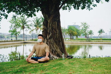 Asian man practicing meditation with wear protective mask on face, yoga exercise under big tree in...