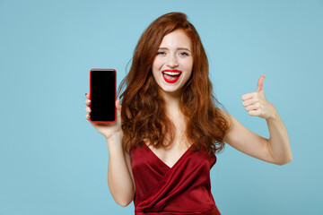 Naklejka premium Young caucasian happy readhead curly woman in red party dress gown hold mobile cell phone with blank screen workspace area show thumb like gesture isolated on pastel blue background studio portrait.