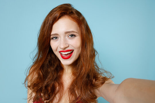 Close Up Young Caucasian Smiling Readhead Curly Woman In Red Party Evening Dress Gown Doing Selfie Shot On Mobile Phone Talking By Video Call Isolated On Pastel Blue Color Background Studio Portrait.