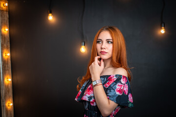 beautiful striking woman with red hair is standing in a black dress with a floral print, standing on a black background with glowing light bulbs. Attractive girl with evening makeup looks away