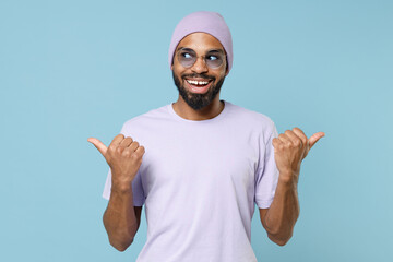 Naklejka premium Young smiling unshaven black dark-skinned african american man 20s in violet t-shirt hat glasses point thumb fingers aside on workspace area mock up isolated on pastel blue background studio portrait.