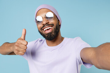 Naklejka premium Close up young happy smiling black fun african man 20s wear violet t-shirt hat glasses doing selfie shot on mobile phone show thumb up like gesture isolated on pastel blue background studio portrait.