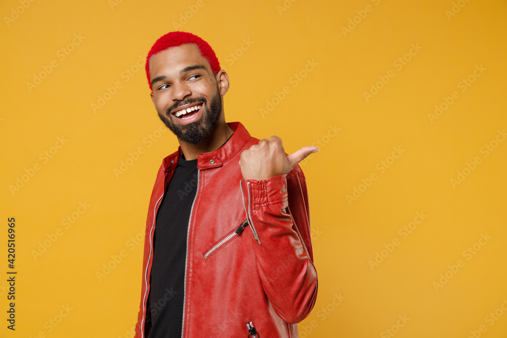 Canvas Prints Young african dark-skinned smiling man 20s with funky trendy pink hairdo wear red leather jacket point thumb finger back aside on workspace area isolated on yellow orange background studio portrait