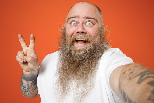 Close up fat pudge obese chubby overweight tattooed blue-eyed bearded man in white tshirt do selfie shot on mobile phone show thumb up like gesture isolated on red orange background studio portrait.