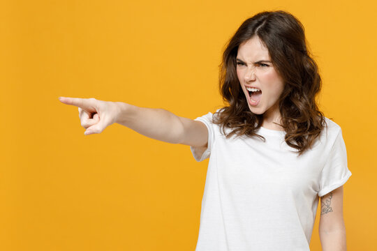 Young strict angry caucasian student woman 20s in white basic casual blank print design t-shirt point index finger camera on you command do it isolated on yellow orange background studio portrait.