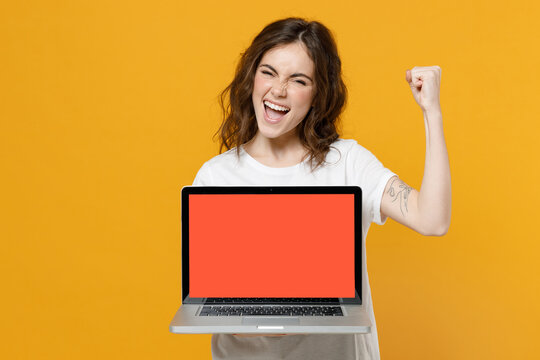 Young overjoyed freelancer student woman in white basic t-shirt hold laptop pc computer with blank screen workspace area doo winner gesture clench fist celebrate isolated on yellow background studio.
